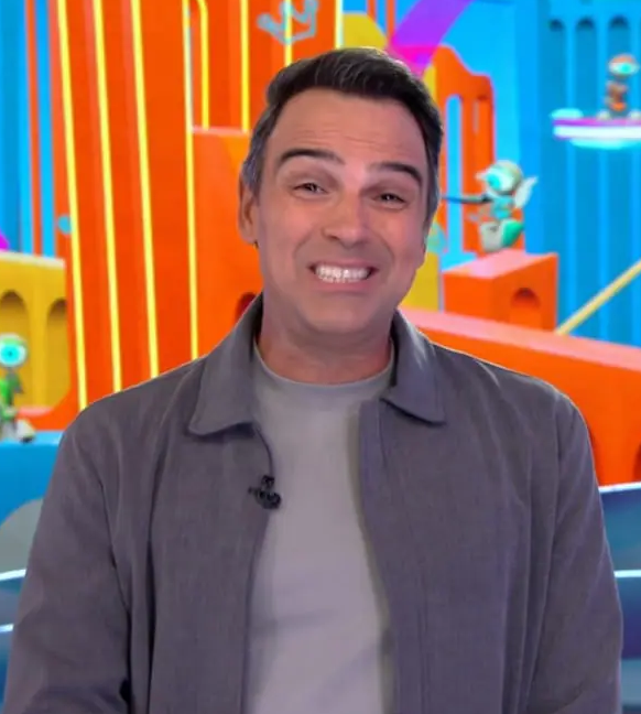 Apresentador Luciano Huck sorri para a câmera em um cenário colorido com elementos lúdicos e robôs ao fundo.
